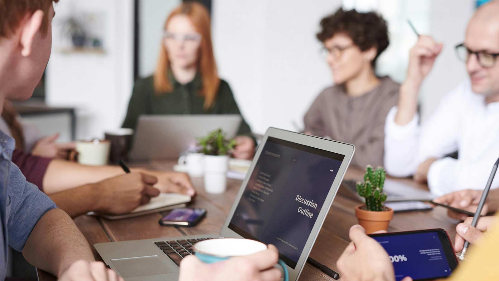 Five people at a meeting table with laptops and a presentation outline on screen