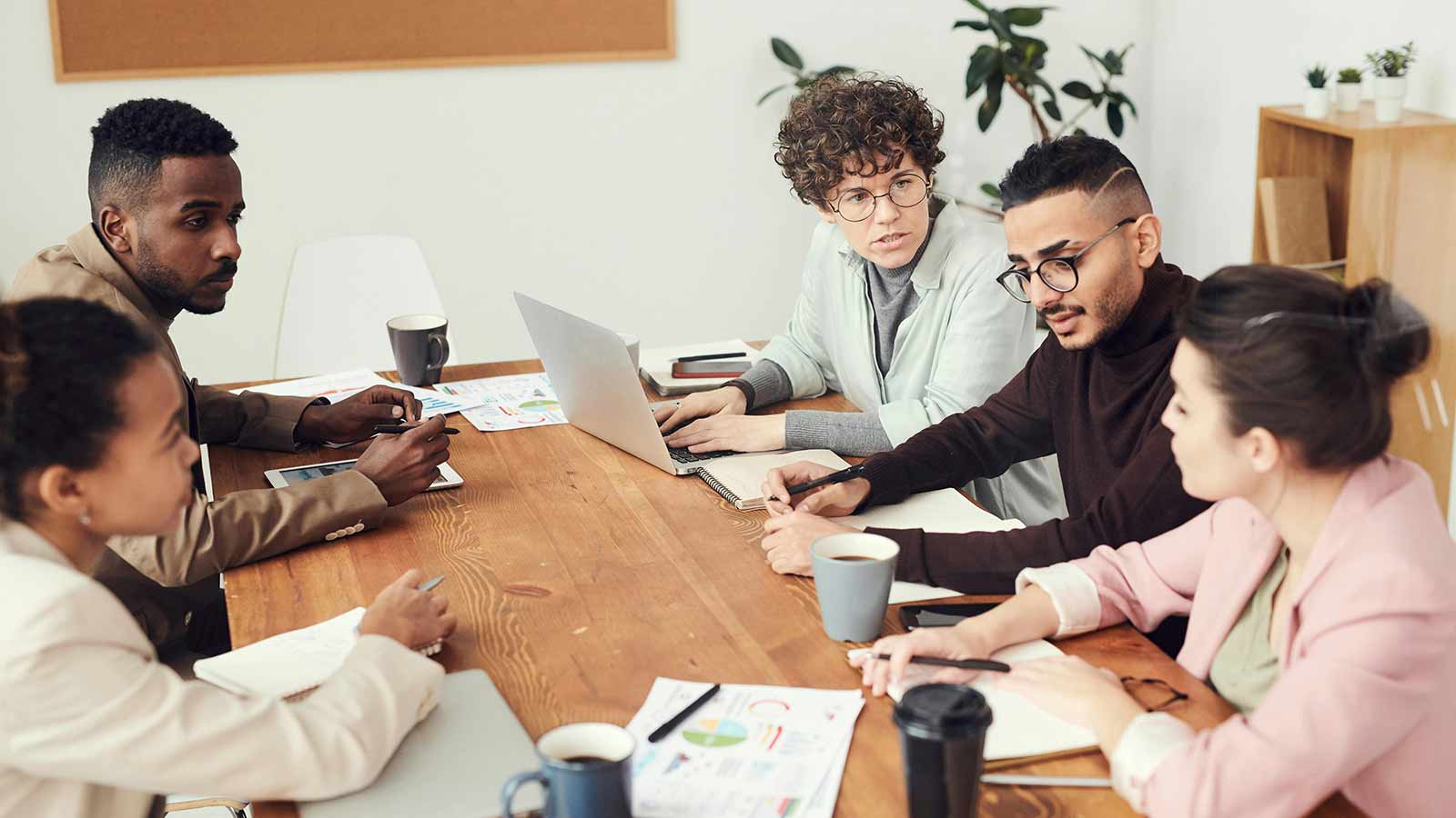 Six colleagues around a wooden conference table with laptops and printed charts