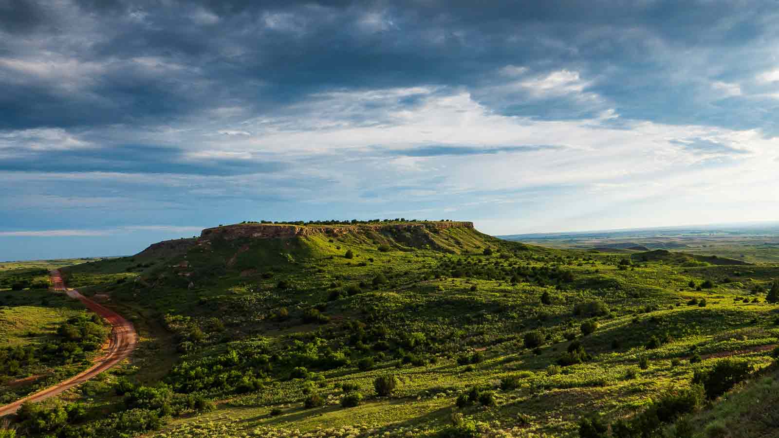 A flat topped mesa rising over green prairie with a red dirt road winding below
